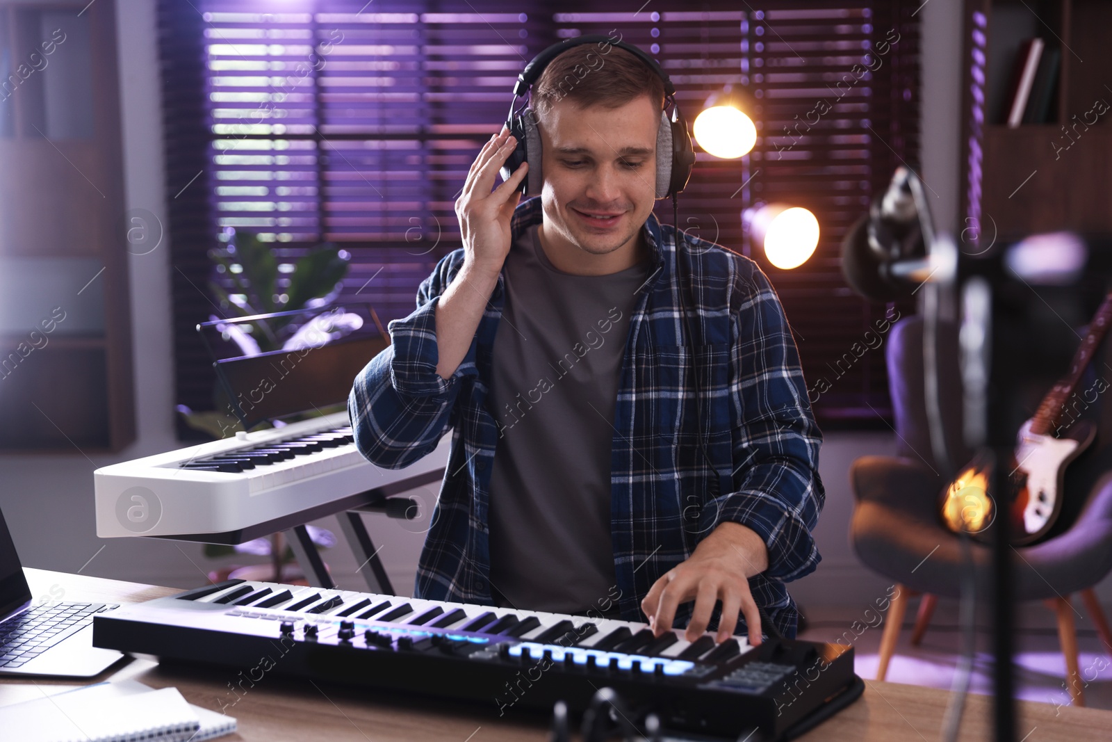 Photo of Smiling man in headphones working with MIDI keyboard at table in home studio