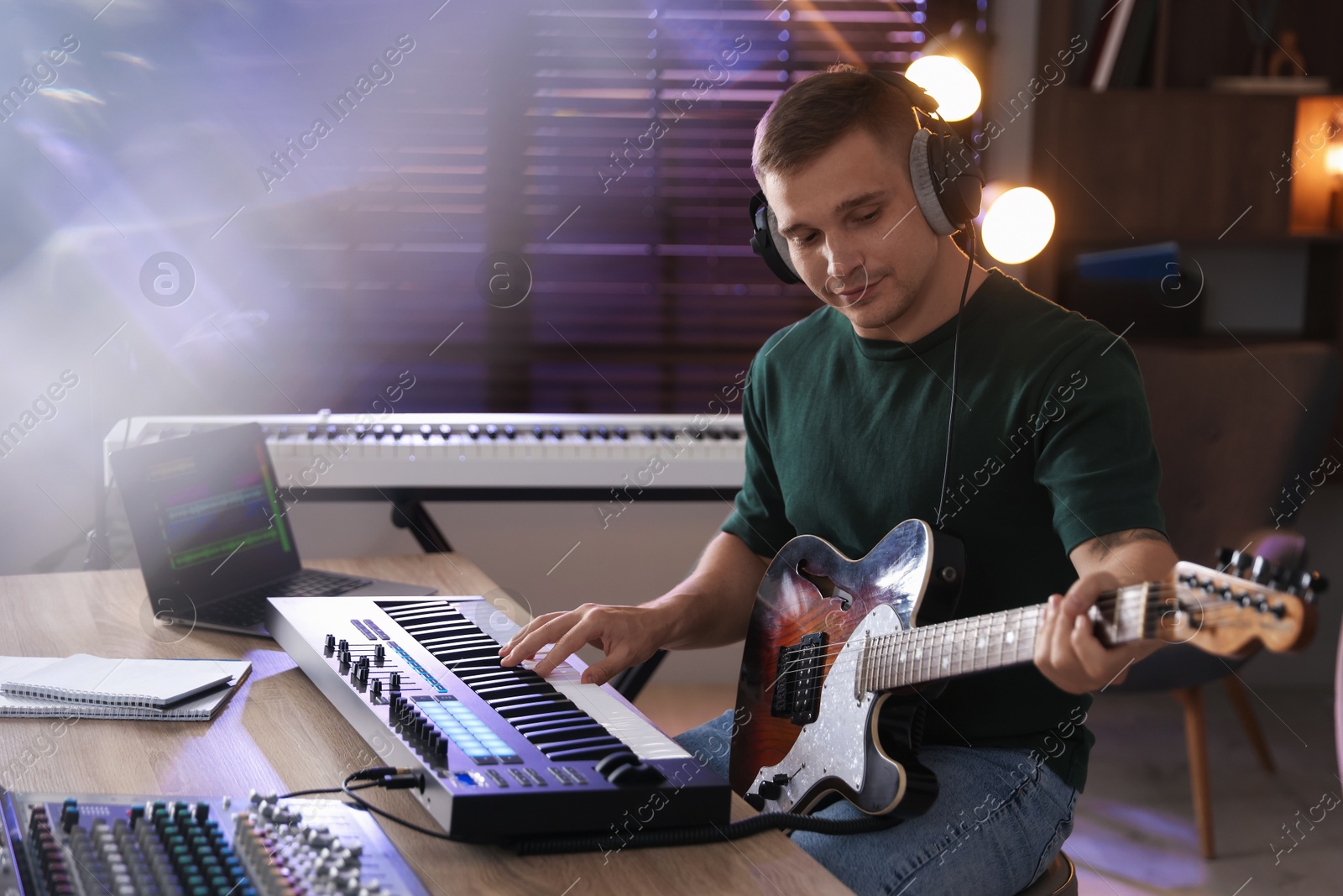 Photo of Man in headphones working with MIDI keyboard and guitar at table in home studio
