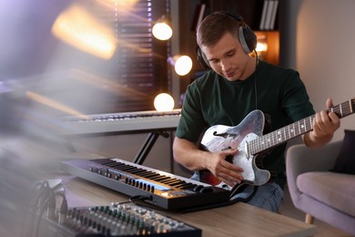 Photo of Man in headphones working with MIDI keyboard and guitar at table in home studio