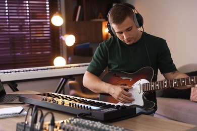 Photo of Man in headphones working with MIDI keyboard and guitar at table in home studio