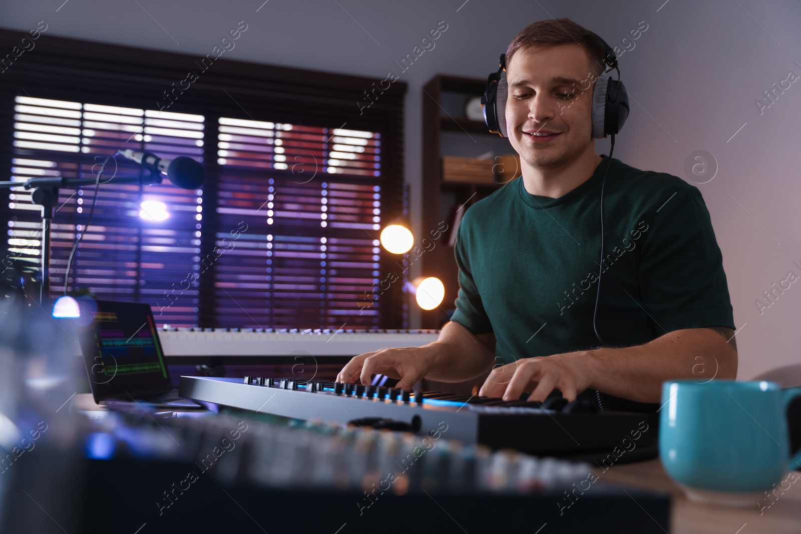 Smiling man in headphones working with MIDI keyboard at table in home studio, low angle view Photo of Smiling man in headphones working with MIDI keyboard at table in home studio, low angle view