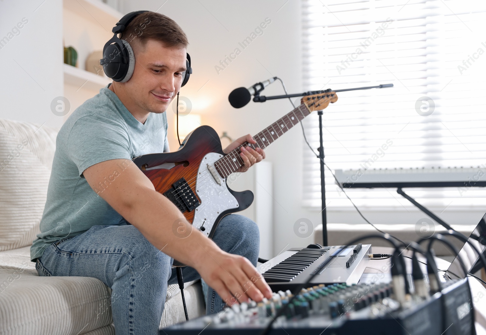 Photo of Man in headphones working with guitar, dj mixer and MIDI keyboard on sofa in home studio, selective focus