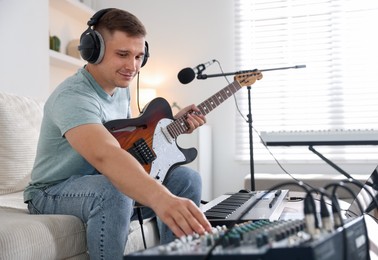 Photo of Man in headphones working with guitar, dj mixer and MIDI keyboard on sofa in home studio, selective focus