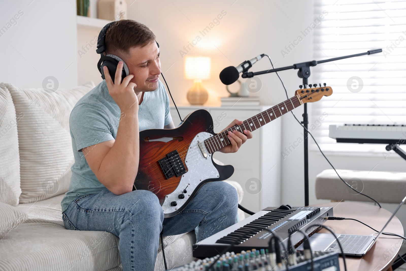Photo of Man in headphones working with guitar and MIDI keyboard on sofa in home studio