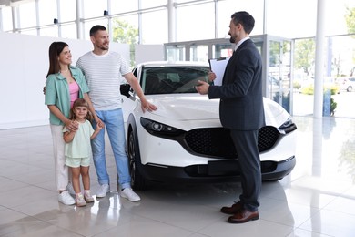 Photo of Salesman with clipboard showing new car to happy family in salon