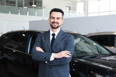 Photo of Portrait of smiling salesman with crossed arms near new car in salon