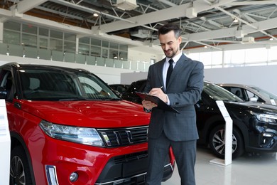 Photo of Smiling salesman working with clipboard near new car in salon