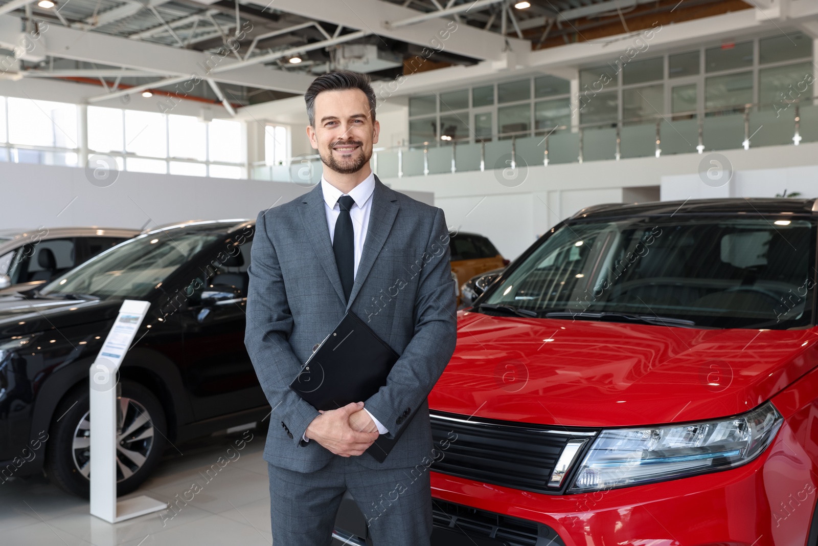 Photo of Portrait of smiling salesman with clipboard near new car in salon