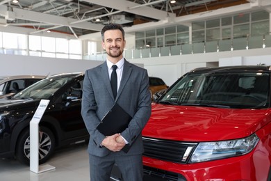 Photo of Portrait of smiling salesman with clipboard near new car in salon