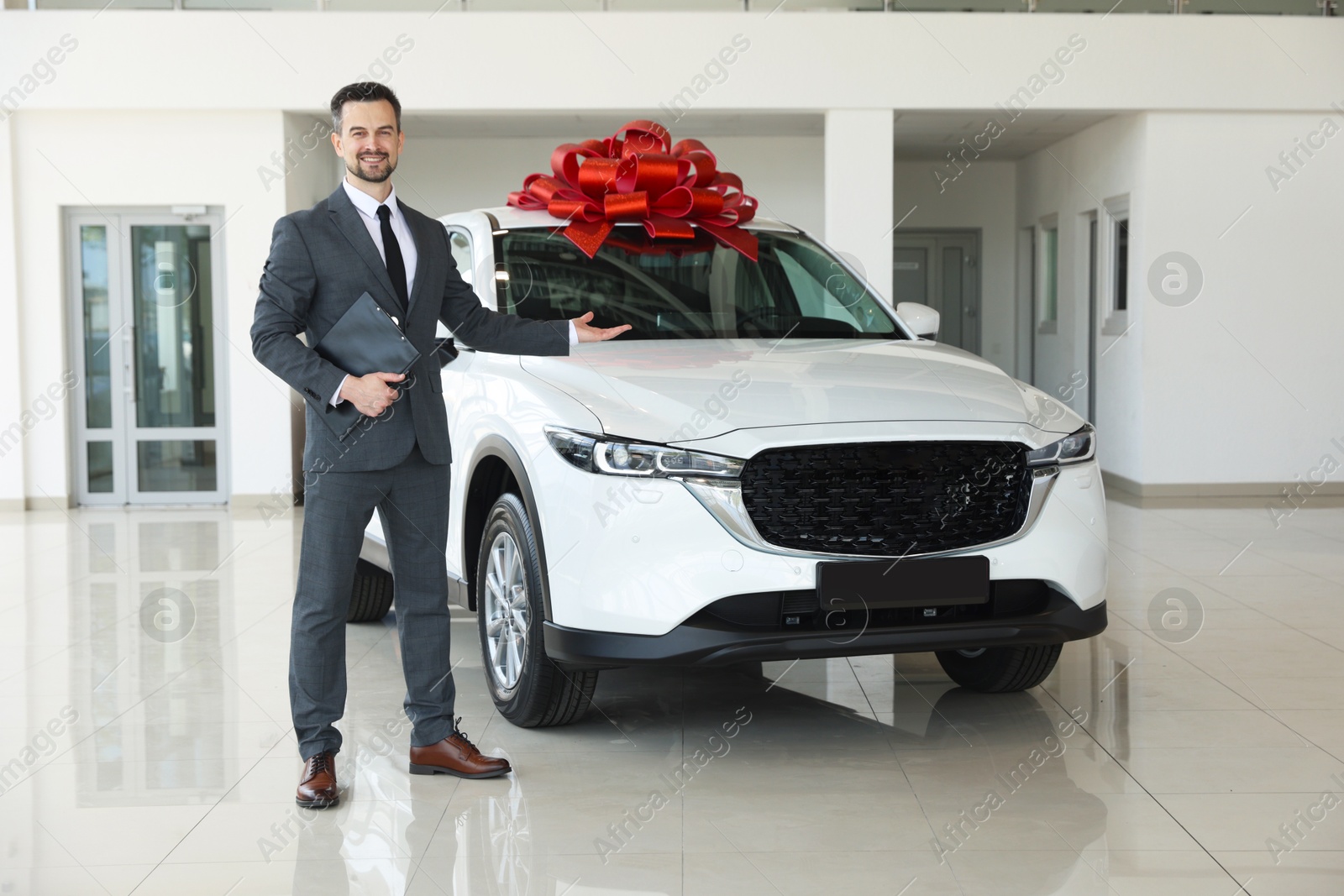 Photo of Smiling salesman with clipboard showing new car in salon