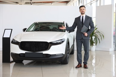 Photo of Smiling salesman with clipboard showing new car in salon