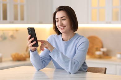 Photo of Woman using modern smartphone at table in kitchen
