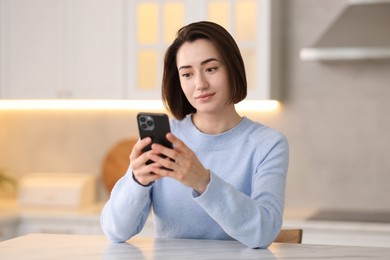 Photo of Woman using modern smartphone at table in kitchen