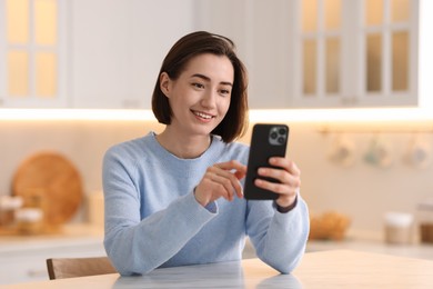 Photo of Woman using modern smartphone at table in kitchen