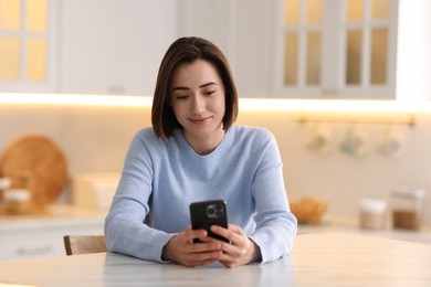 Photo of Woman using modern smartphone at table in kitchen