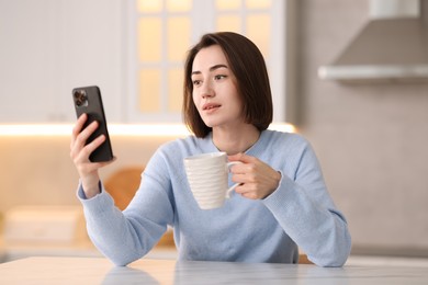 Photo of Woman with cup of drink using modern smartphone at table in kitchen