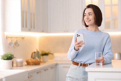 Photo of Woman with glass of water using modern smartphone at table in kitchen. Space for text