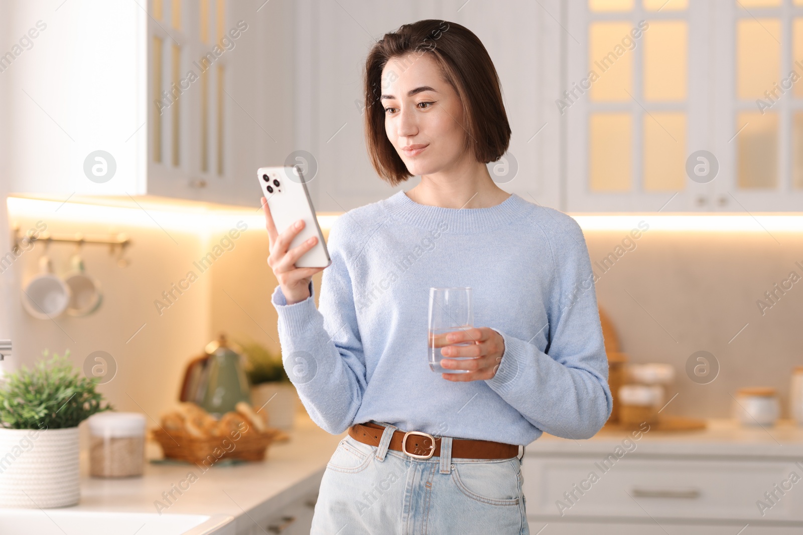 Photo of Woman with glass of water using modern smartphone in kitchen