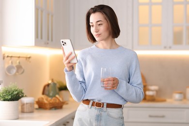 Photo of Woman with glass of water using modern smartphone in kitchen