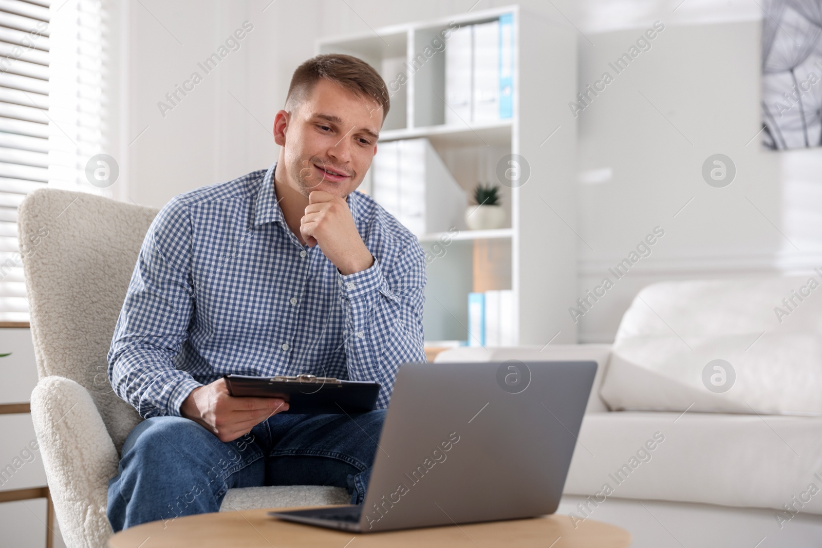 Photo of Smiling psychologist with clipboard during online session by laptop in office