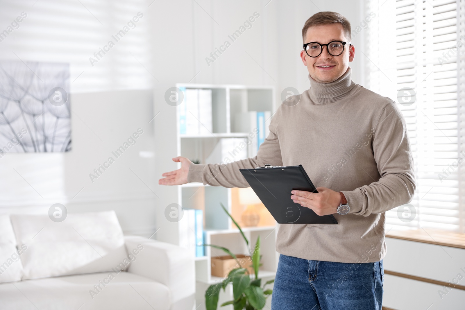 Photo of Smiling psychologist in glasses with clipboard inviting to sofa in office