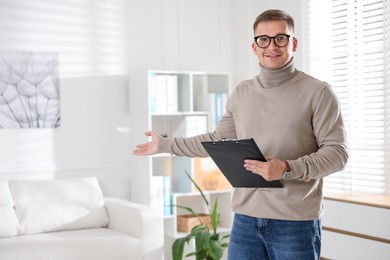 Photo of Smiling psychologist in glasses with clipboard inviting to sofa in office