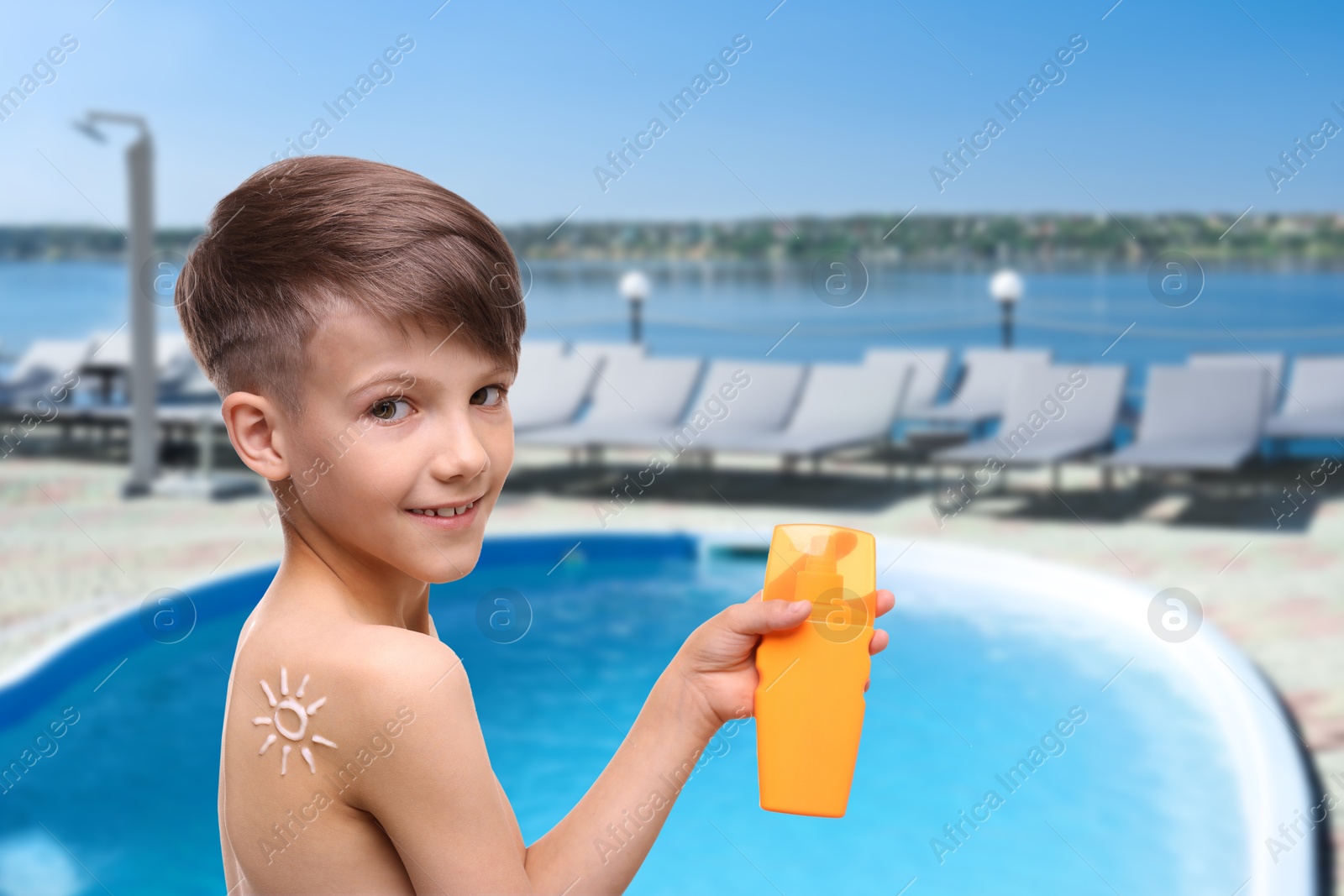 Image of Little boy with bottle of sunscreen by outdoor swimming pool. Sun protection product on his shoulder