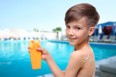 Image of Little boy with bottle of sunscreen by outdoor swimming pool. Sun protection product on his shoulder