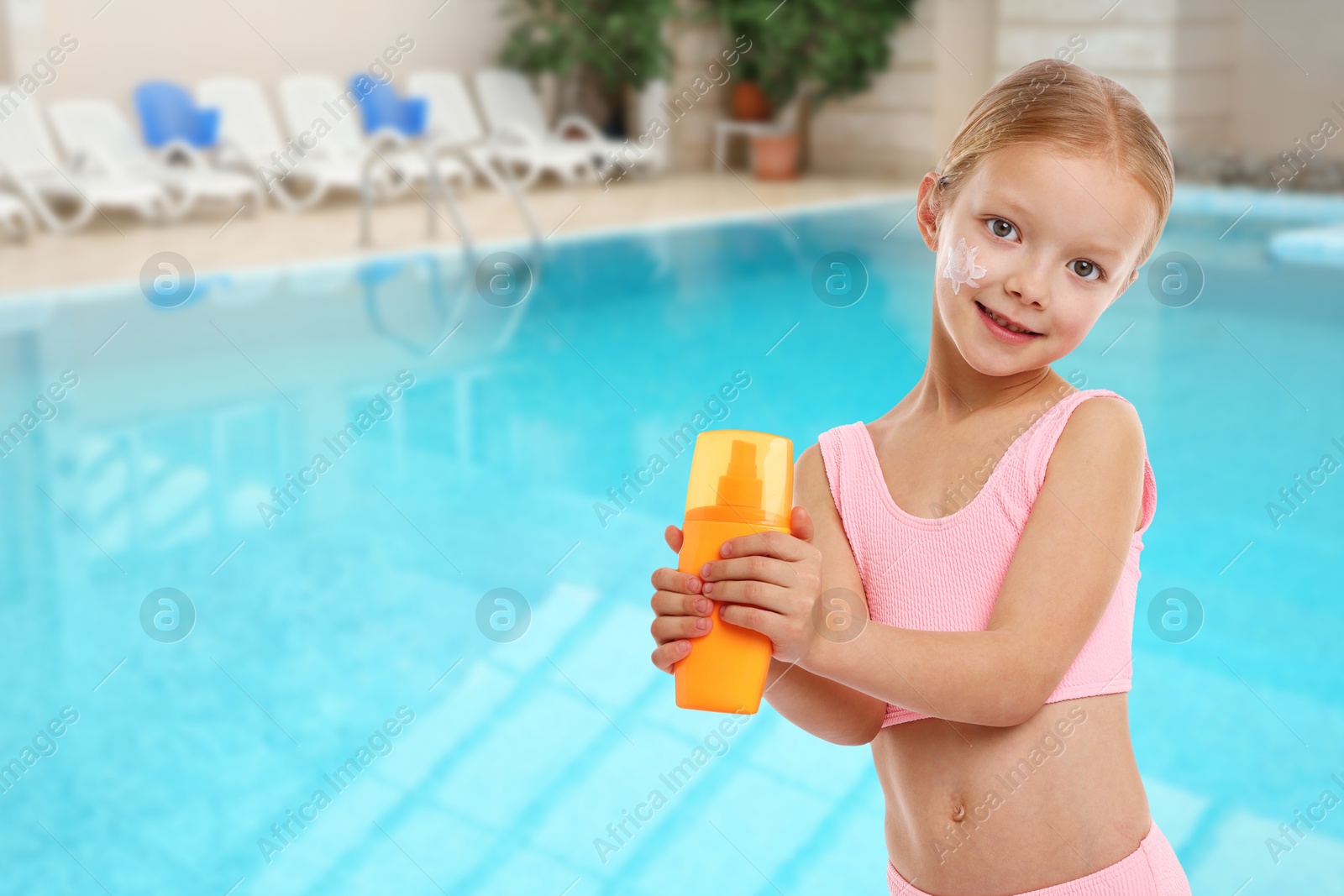 Image of Cute little girl with bottle of sunscreen by outdoor swimming pool. Sun protection product on her cheek