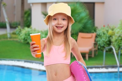 Image of Cute little girl with tube of sunscreen and inflatable ring by outdoor swimming pool. Sun protection product on her cheek