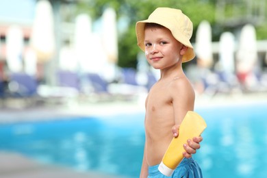 Image of Little boy with tube of sunscreen by outdoor swimming pool. Sun protection product on his skin