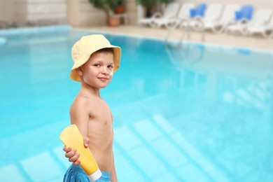 Image of Little boy with tube of sunscreen by outdoor swimming pool. Sun protection product on his skin