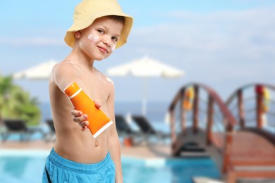 Image of Little boy with tube of sunscreen by outdoor swimming pool. Sun protection product on his skin