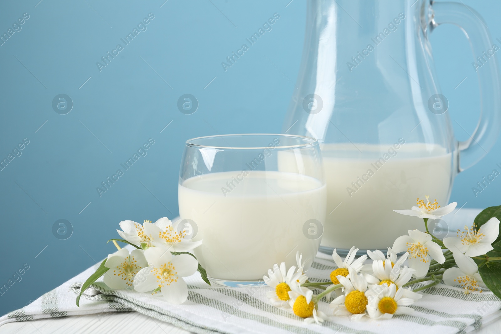 Photo of Fresh milk and flowers on white wooden table against light blue background, closeup. Dairy product