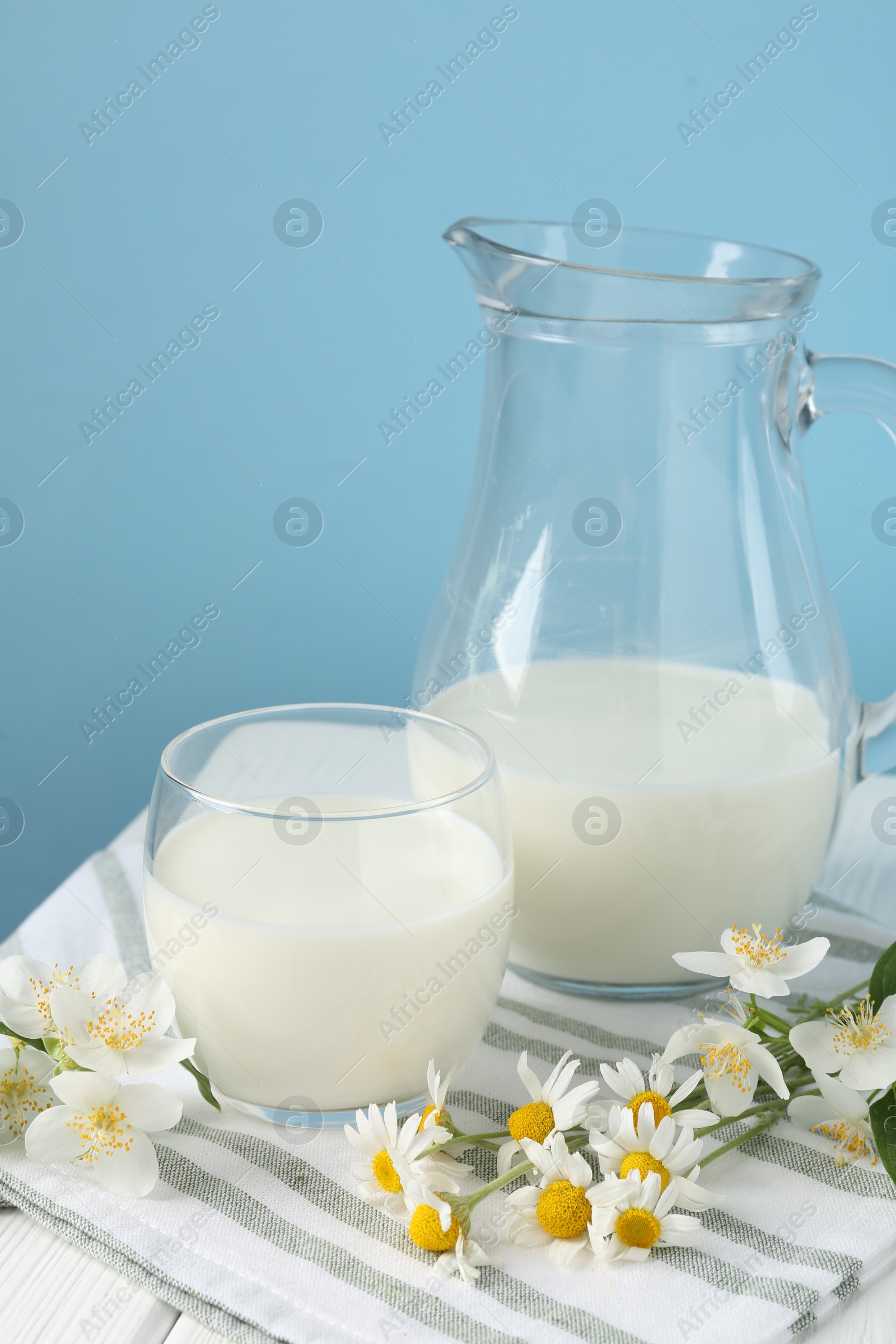 Photo of Fresh milk and flowers on white wooden table against light blue background, closeup. Dairy product