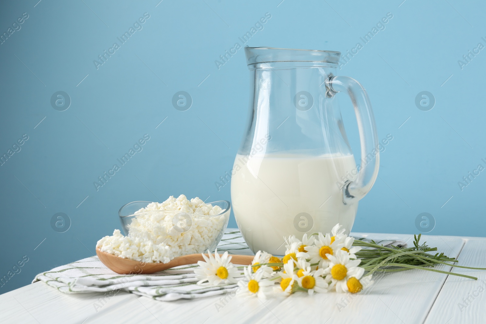 Photo of Fresh milk in jug, cottage cheese and chamomile flowers on white wooden table against light blue background. Dairy products