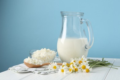 Photo of Fresh milk in jug, cottage cheese and chamomile flowers on white wooden table against light blue background. Dairy products