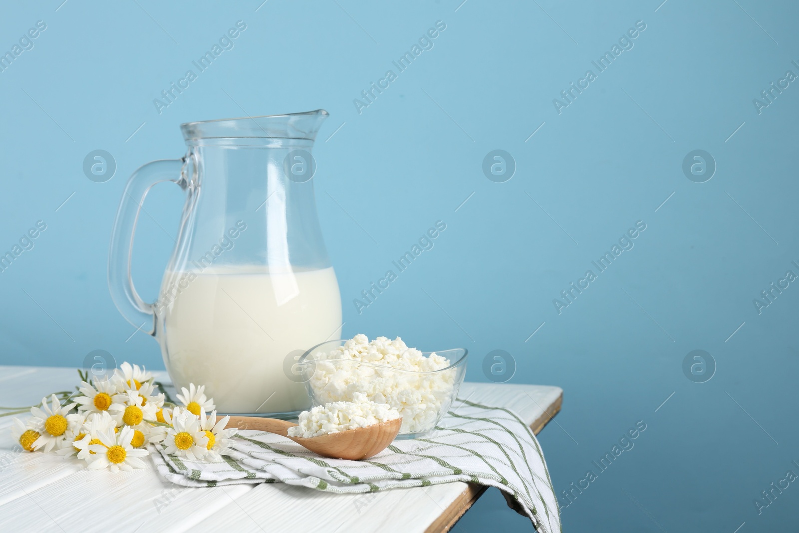 Photo of Fresh milk in jug, cottage cheese and chamomile flowers on white wooden table against light blue background, space for text. Dairy products