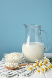 Photo of Fresh milk in jug, cottage cheese and chamomile flowers on white wooden table against light blue background. Dairy products