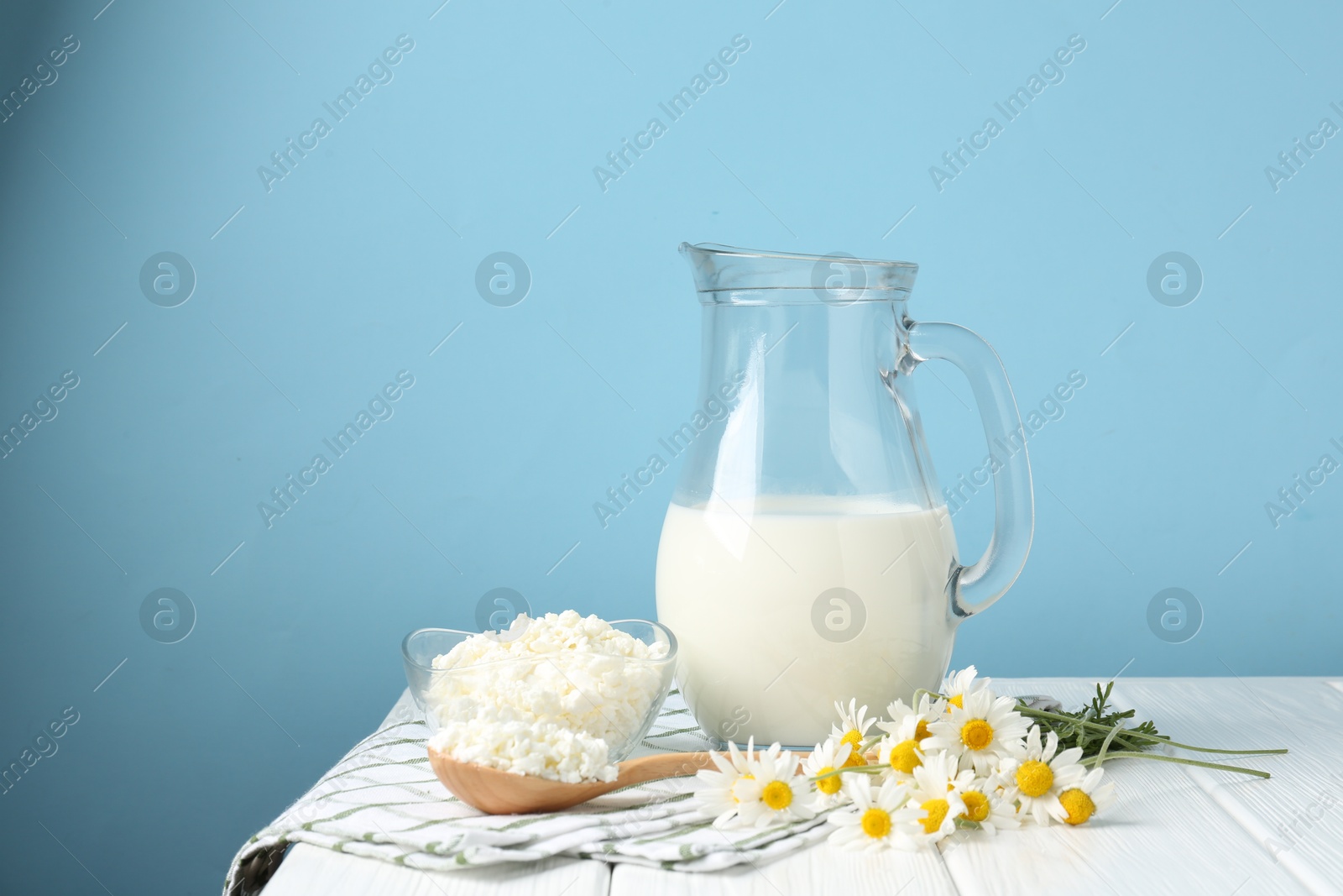 Photo of Fresh milk in jug, cottage cheese and chamomile flowers on white wooden table against light blue background, space for text. Dairy products