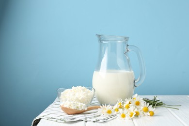 Photo of Fresh milk in jug, cottage cheese and chamomile flowers on white wooden table against light blue background, space for text. Dairy products