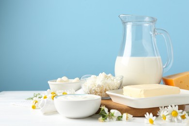 Photo of Fresh dairy products and flowers on white wooden table against light blue background