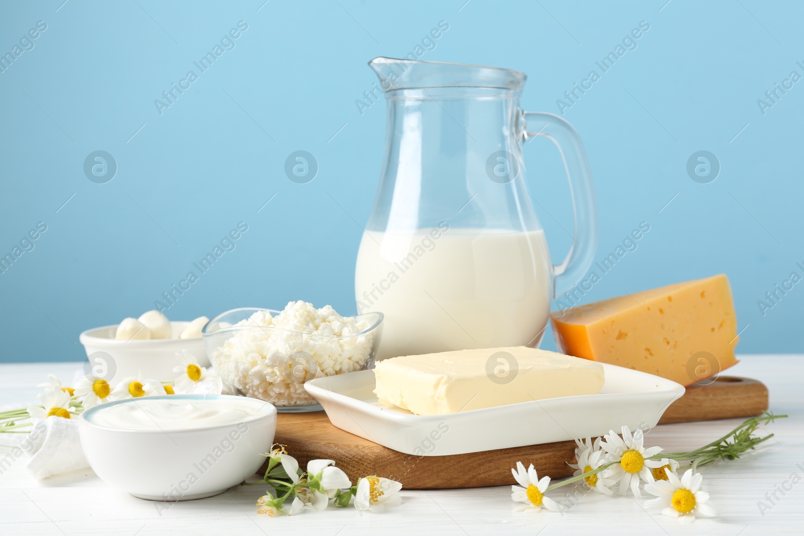 Photo of Fresh dairy products and flowers on white wooden table against light blue background, closeup