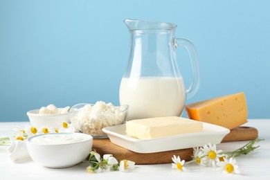 Photo of Fresh dairy products and flowers on white wooden table against light blue background, closeup