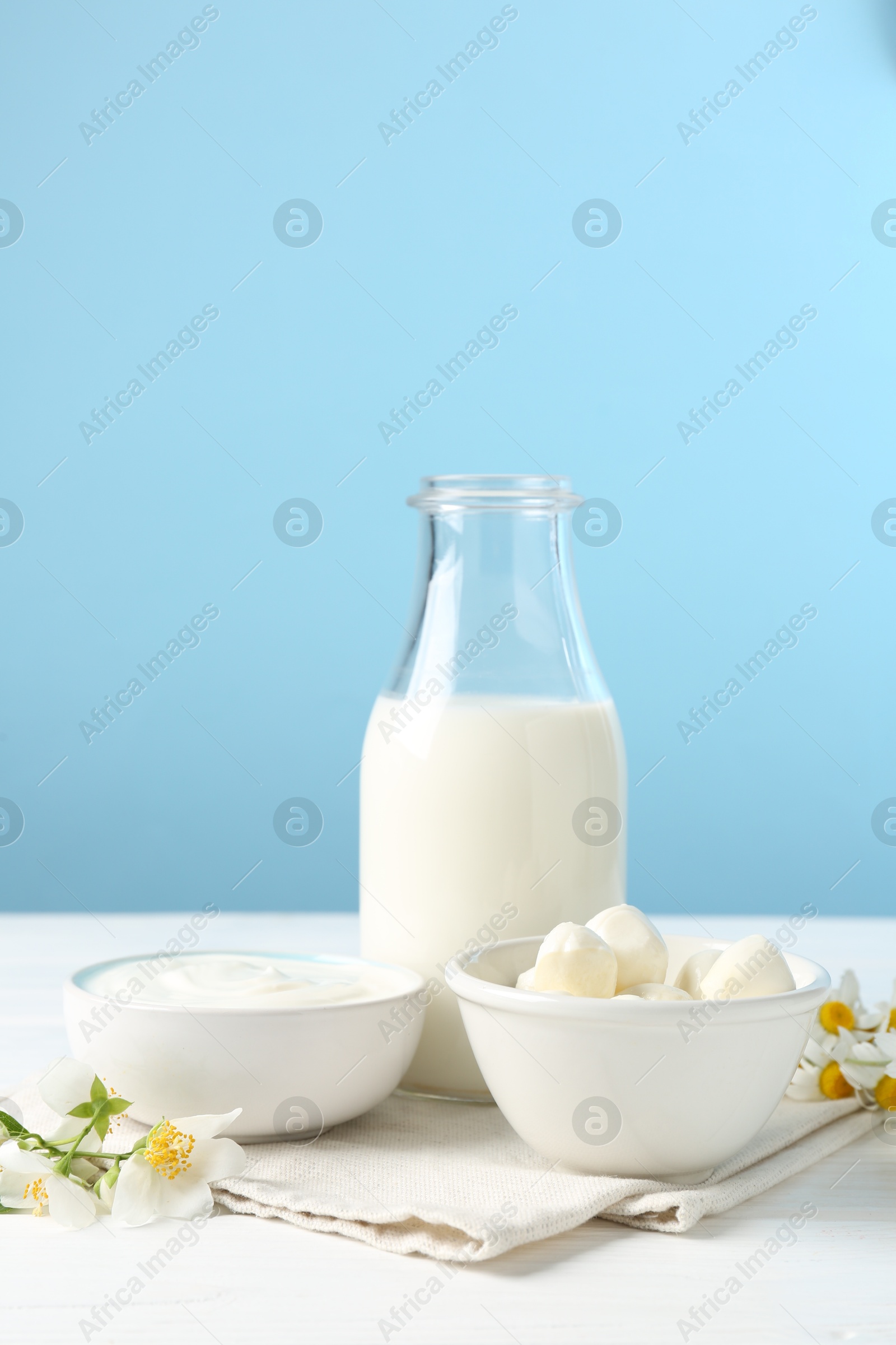 Photo of Fresh dairy products and flowers on white wooden table against light blue background