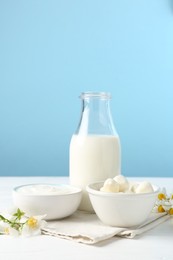 Photo of Fresh dairy products and flowers on white wooden table against light blue background