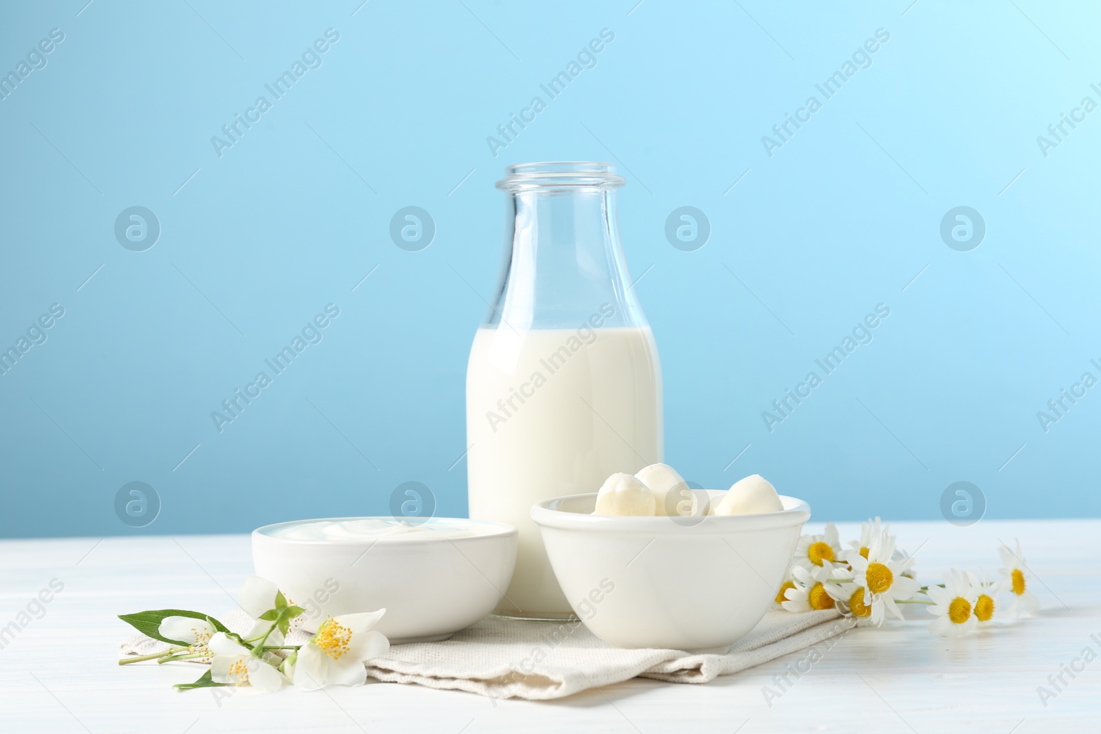 Photo of Fresh dairy products and flowers on white wooden table against light blue background