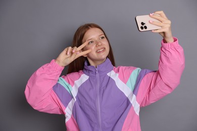 Photo of Smiling teenage girl taking selfie by smartphone and showing peace sign on grey background