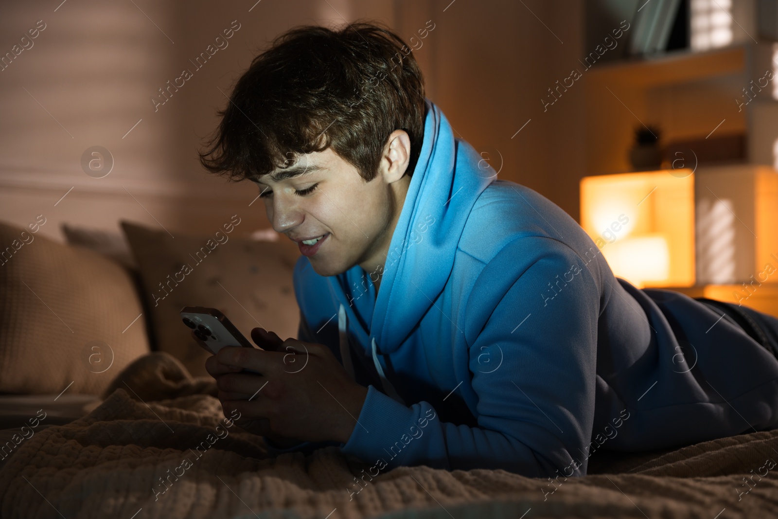 Photo of Smiling teenage boy using smartphone on bed at home