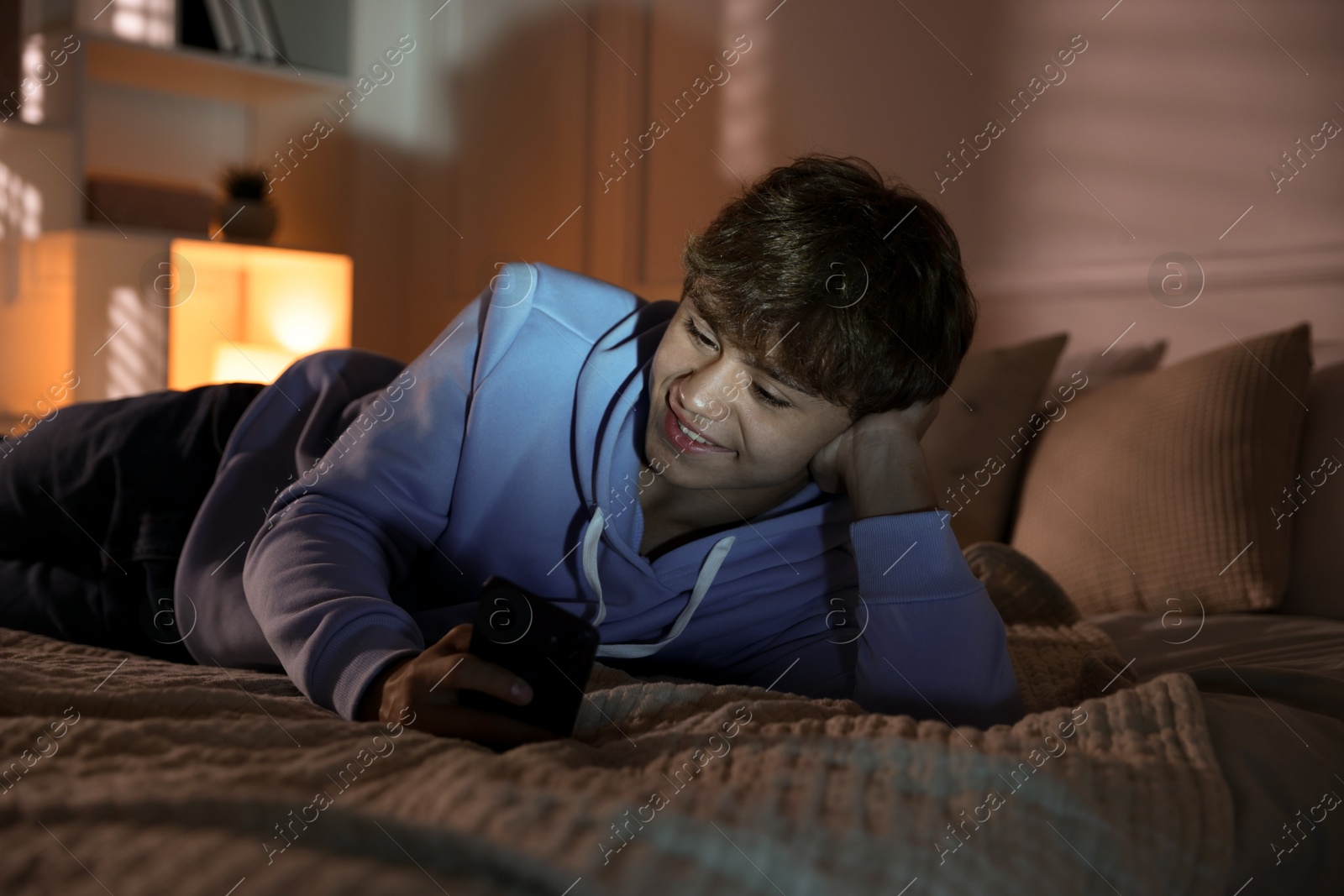 Photo of Smiling teenage boy using smartphone on bed at home